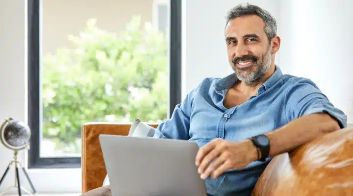 A man sitting at a table with a laptop and smiling at the camera